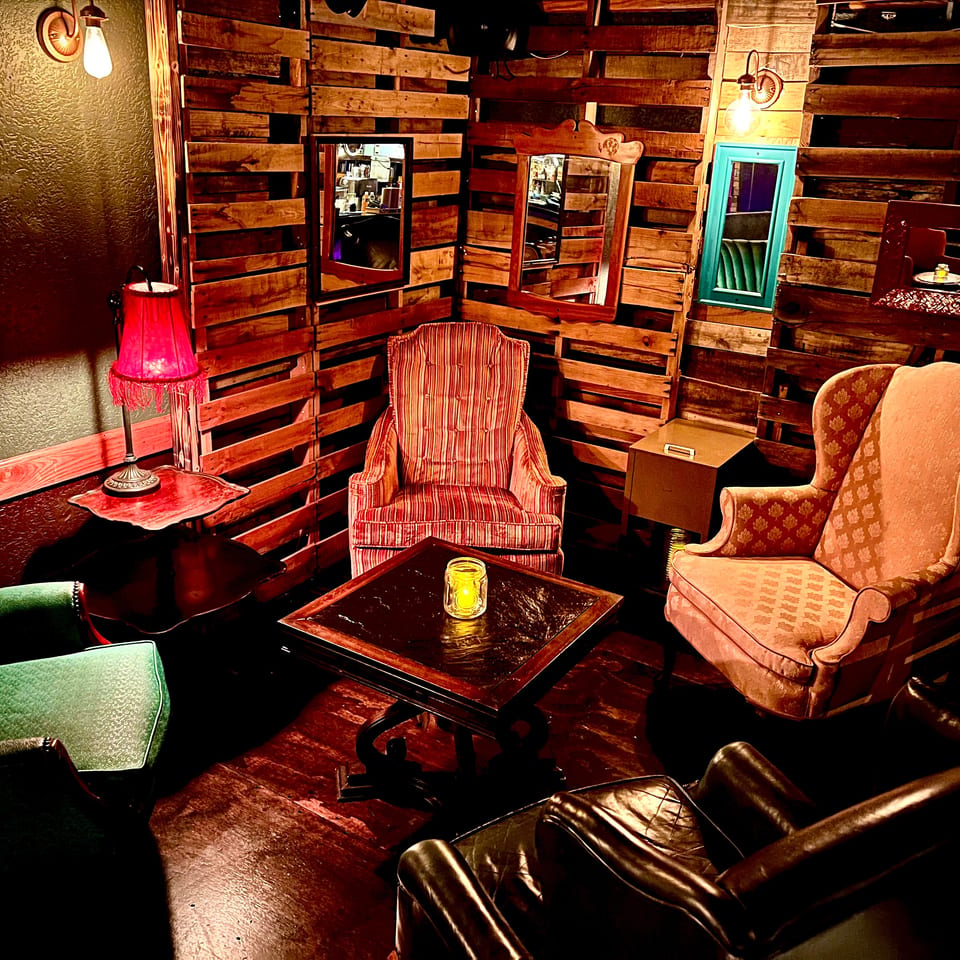 Cozy restaurant lounge with mismatched vintage armchairs, candlelit tables, warm lamps, and rustic wood-plank walls.