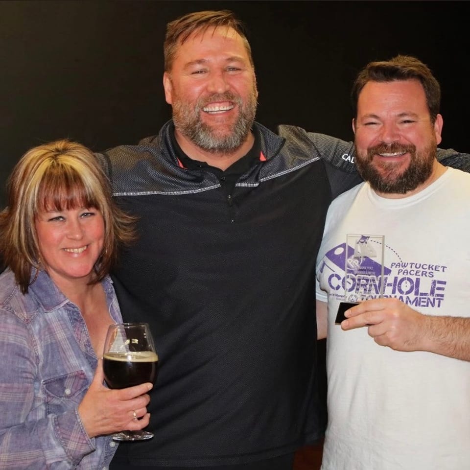 Three smiling people pose together at a bar, with one holding a glass of dark beer and another wearing a cornhole tournament shirt.