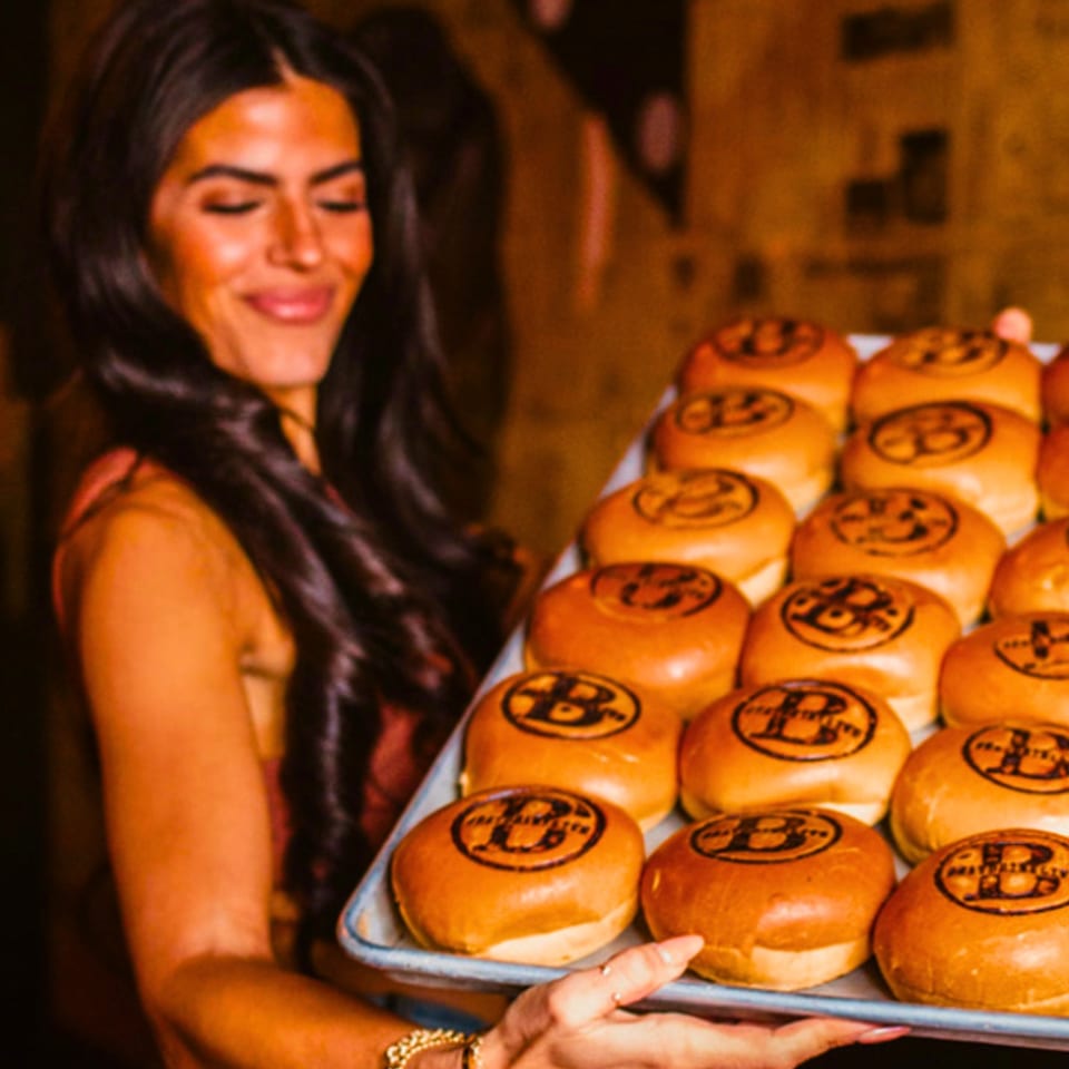 Smiling woman carries a tray of golden pastries stamped with playful designs in a warmly lit restaurant.