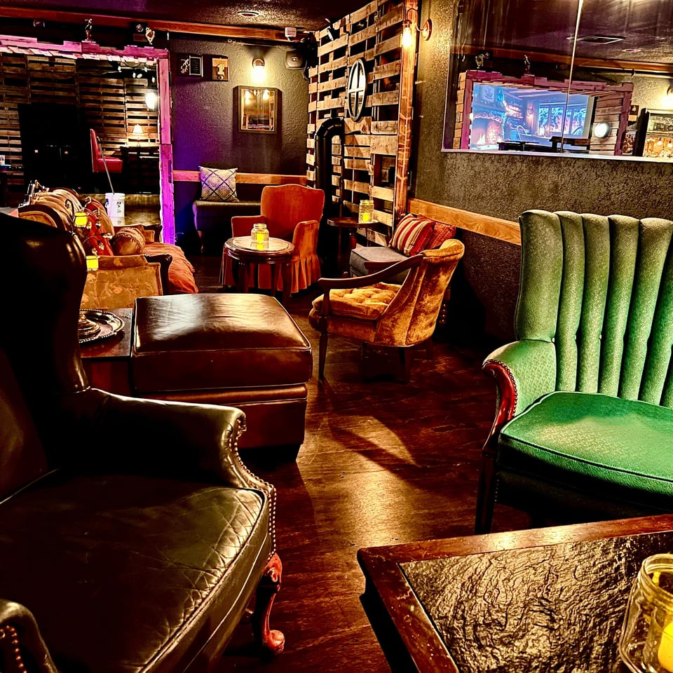 Cozy restaurant lounge with vintage leather seating, colorful upholstered chairs, wooden walls, candlelit tables, and a service window.