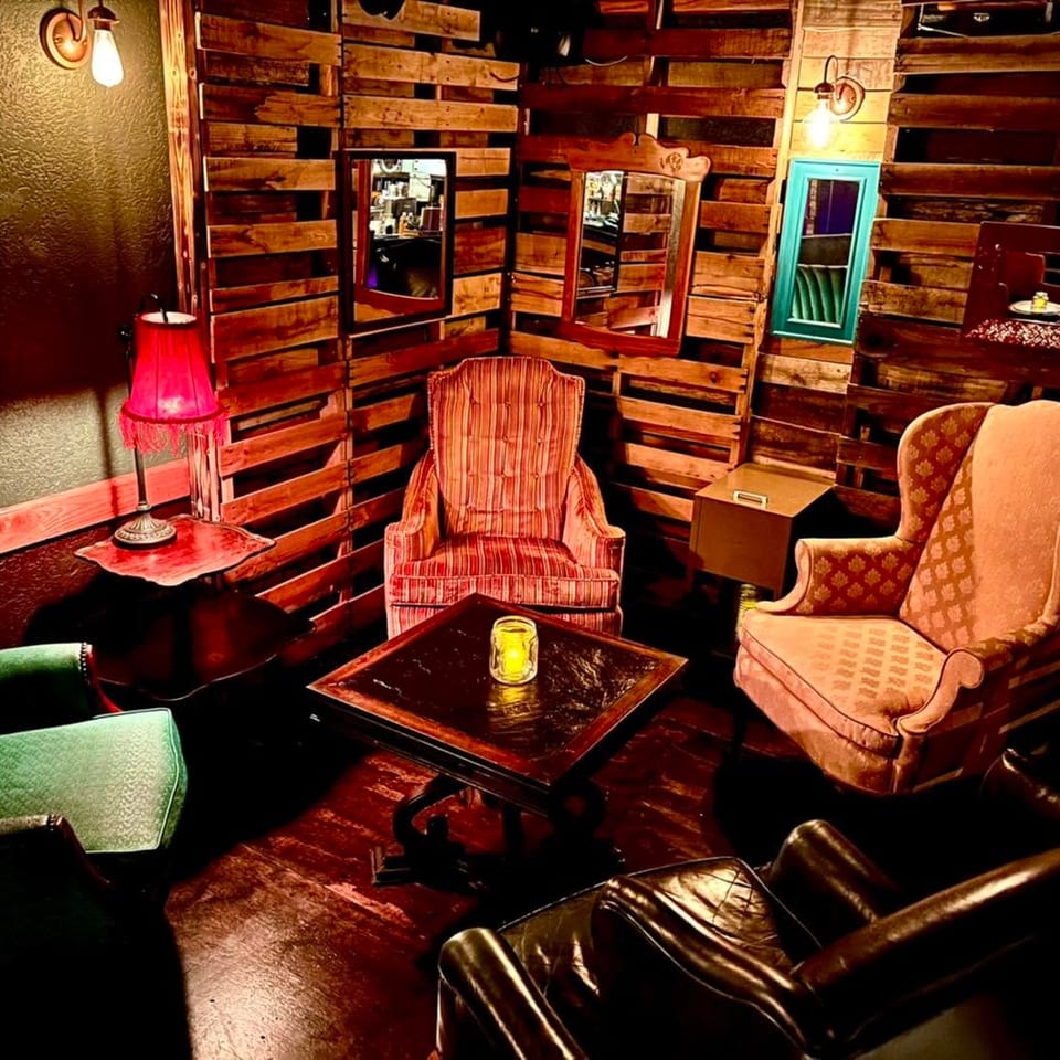 Cozy lounge area with mismatched armchairs, wooden pallet walls, and warm lighting from lamps and candles.