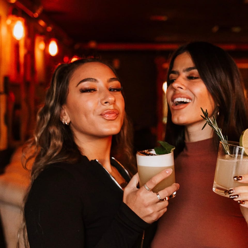 Two women smile with cocktails in a warmly lit restaurant or bar with red ambient lighting.