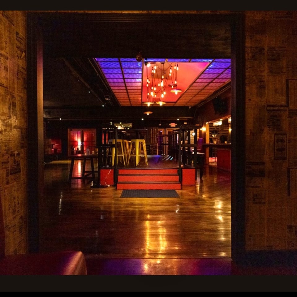 Dimly lit restaurant interior with newspaper-covered walls, polished wood floors, colorful ceiling lights, bar seating, and booths.