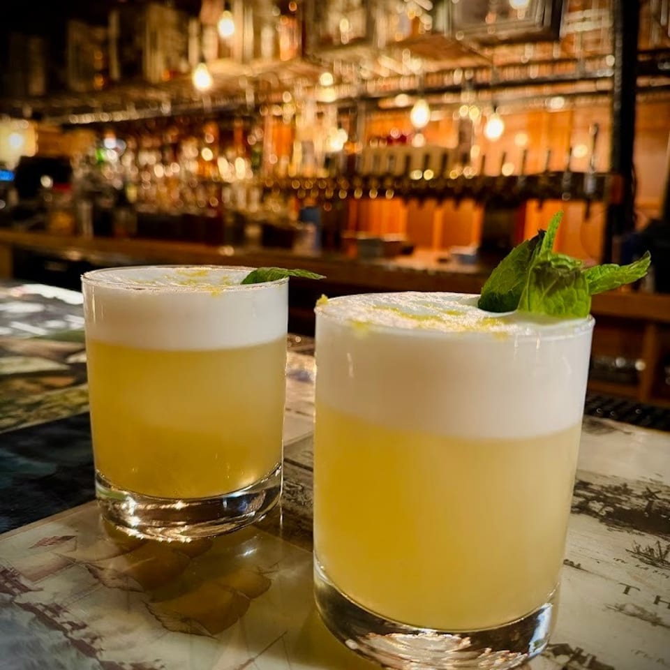 Two frothy yellow cocktails garnished with fresh mint sit on a table in front of a warmly lit bar.