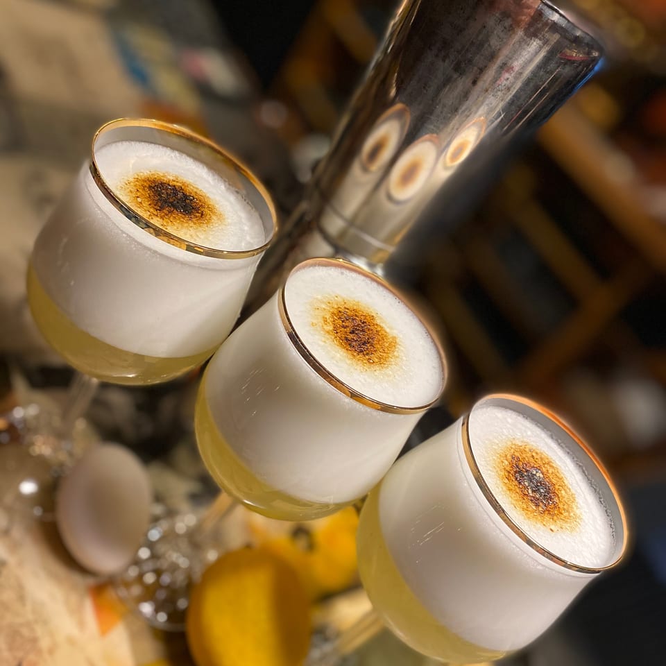 Three gold-rimmed cocktails topped with foam and toasted spice sit among lemons and barware in warm, softly lit surroundings.