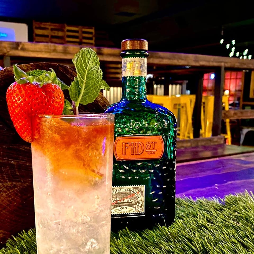 A tall cocktail with a strawberry and mint garnish next to a green bottle of Fid St Gin.