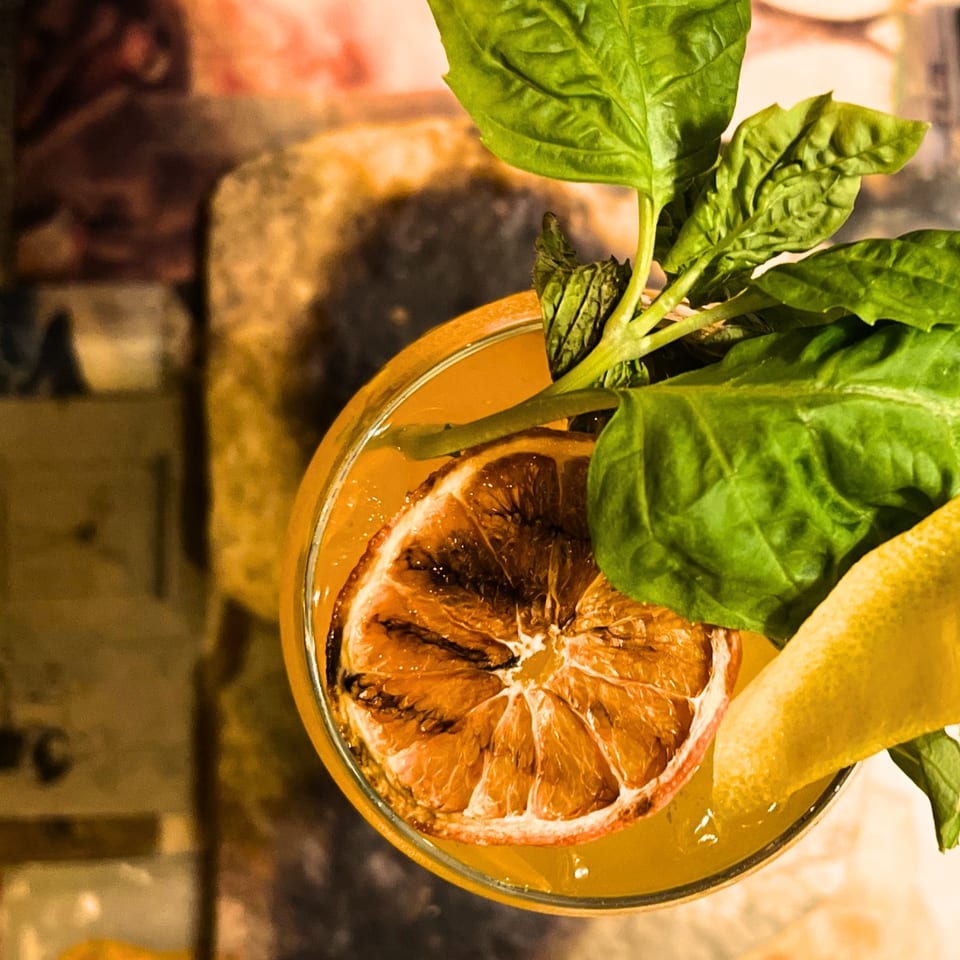 Citrus cocktail garnished with a halved grapefruit, lemon wedge, and fresh leafy herbs on a warmly lit bar.