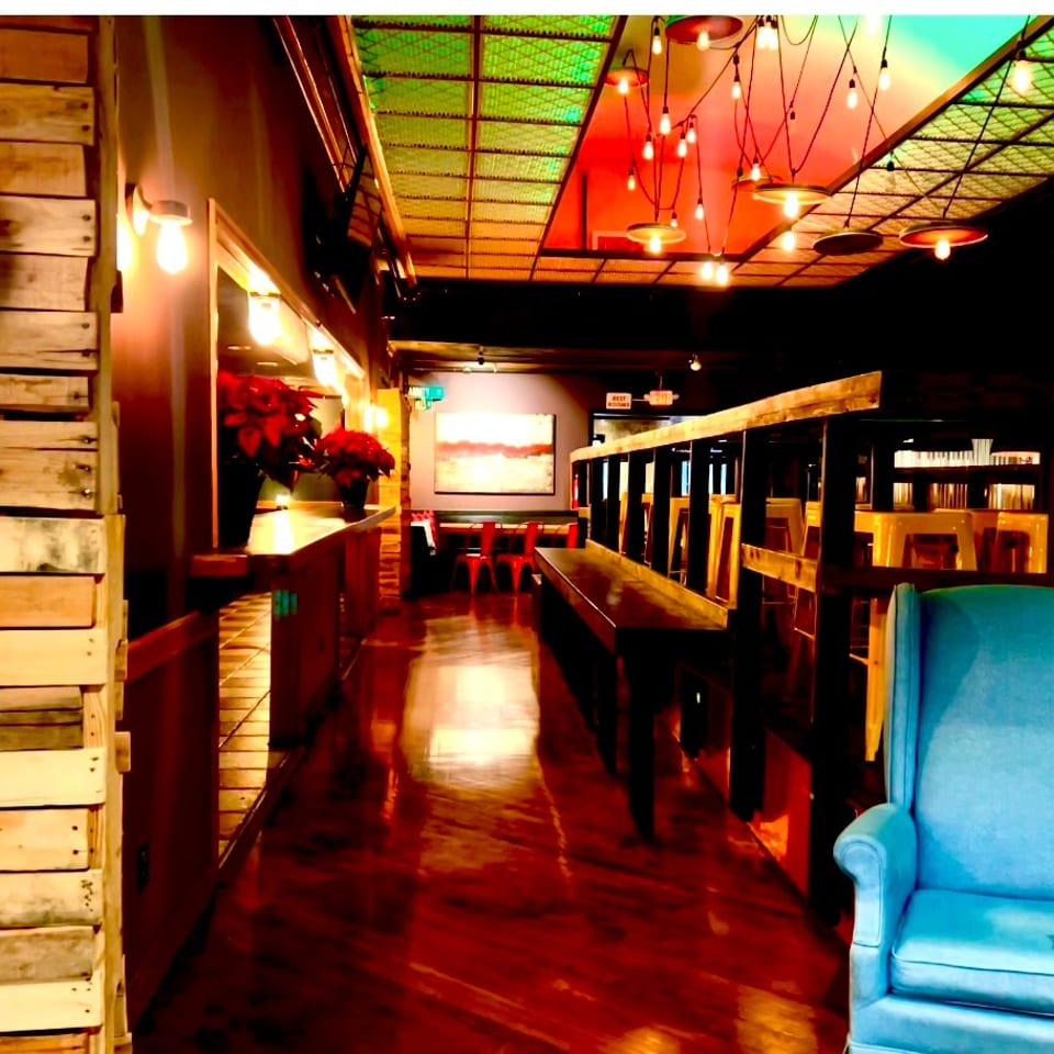 Warmly lit restaurant interior with wood-paneled walls, colorful ceiling lights, booths, bar seating, and a bright blue armchair.