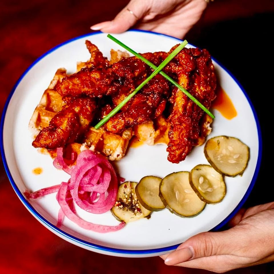 Hands holding a plate of crispy fried chicken and waffles, garnished with pickled onions and cucumbers.