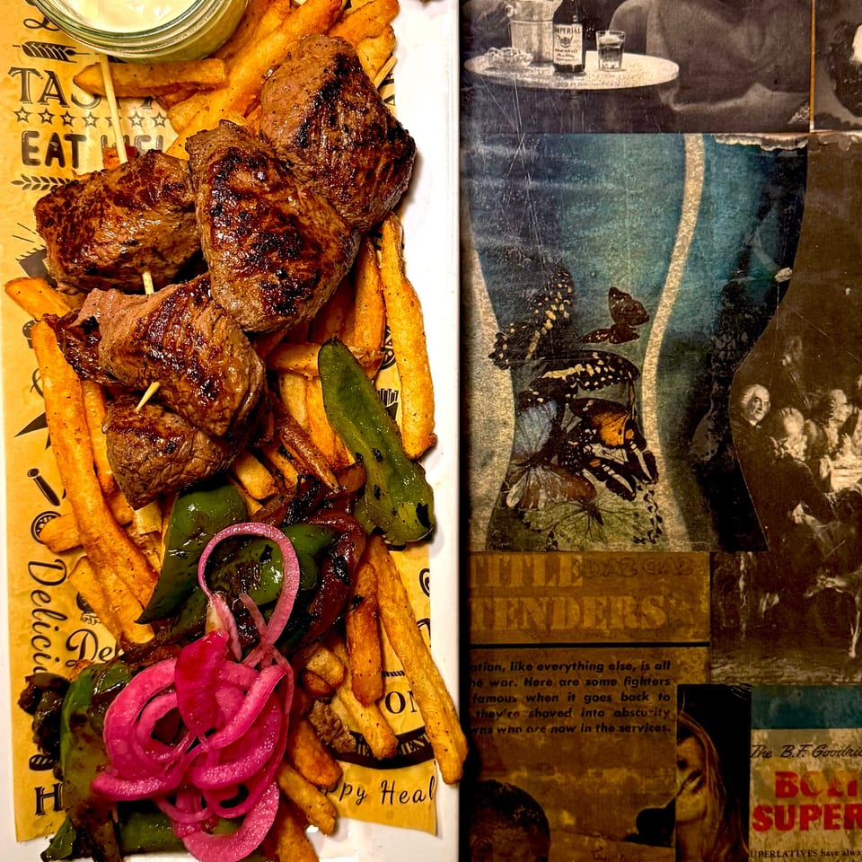 Grilled beef skewers with fries, grilled peppers, pickled onions, and creamy dipping sauce on a white plate beside vintage newspaper décor.