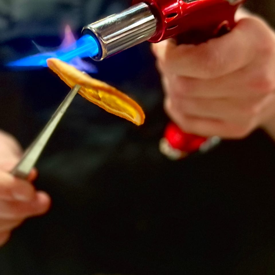 Bartender uses a red torch to caramelize an orange peel over a cocktail.